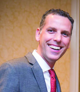 Man smiling, wearing a red tie, white collared shirt and gray suit jacket